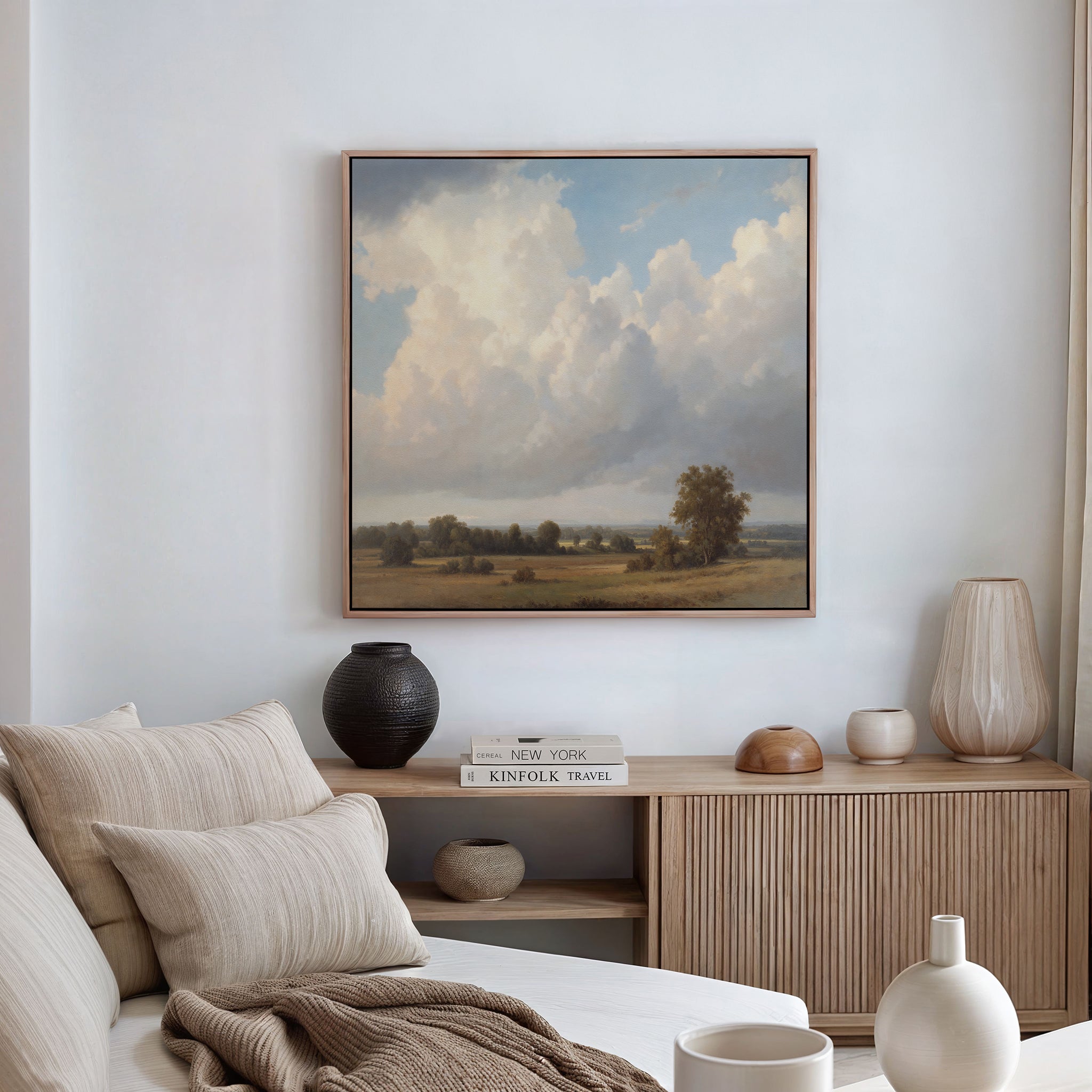 A cozy, neutral-toned living room with a beige sofa, textured cushions, and a wooden sideboard with vases, books, and decor. Above hangs the Pastoral Grandeur Canvas Art - Square, adding serene countryside charm to the space.