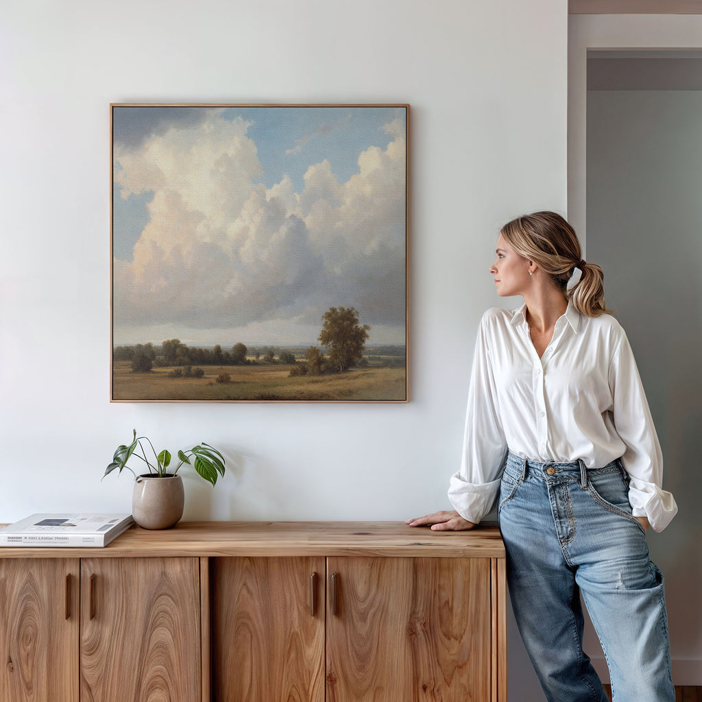 A woman in a white blouse and jeans leans against a wooden cabinet, gazing at the Pastoral Grandeur Canvas Art - Square featuring clouds and fields. A potted plant and books atop the cabinet enhance the tranquil scene.