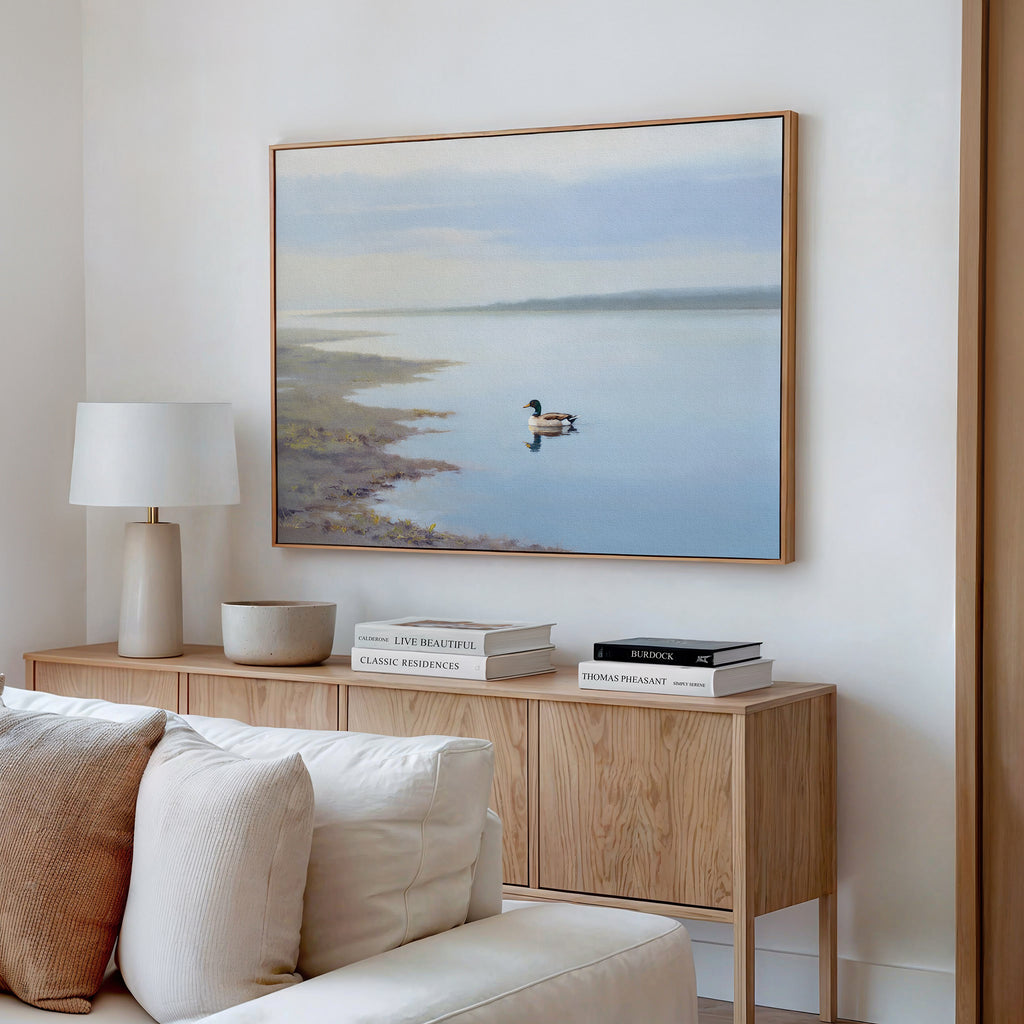 A minimal living room with a light wooden sideboard, books, a lamp, and the Peaceful Duck Haven Canvas Art - Horizontal above a white sofa creates a tranquil Nordic Park vibe.