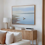 A minimal living room with a light wooden sideboard, books, a lamp, and the Peaceful Duck Haven Canvas Art - Horizontal above a white sofa creates a tranquil Nordic Park vibe.
