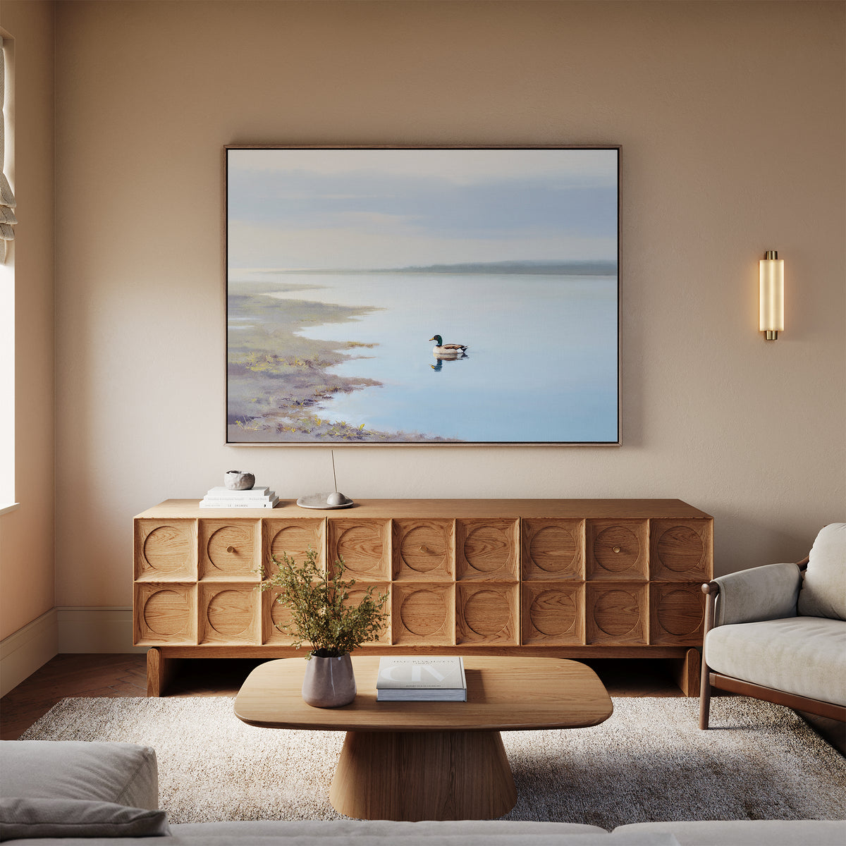 A serene living room with a light wood sideboard, the Peaceful Duck Haven Canvas Art - Horizontal above, a small lamp, a round coffee table with flowers and a book, and a beige sofa on a soft rug.