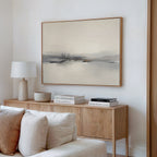 A modern minimalist living room features a light wood sideboard, white lamp, books, and bowl beneath the Peaceful Lake Morning Canvas Art - Horizontal, hanging above a beige couch with cushions to create a peaceful atmosphere.