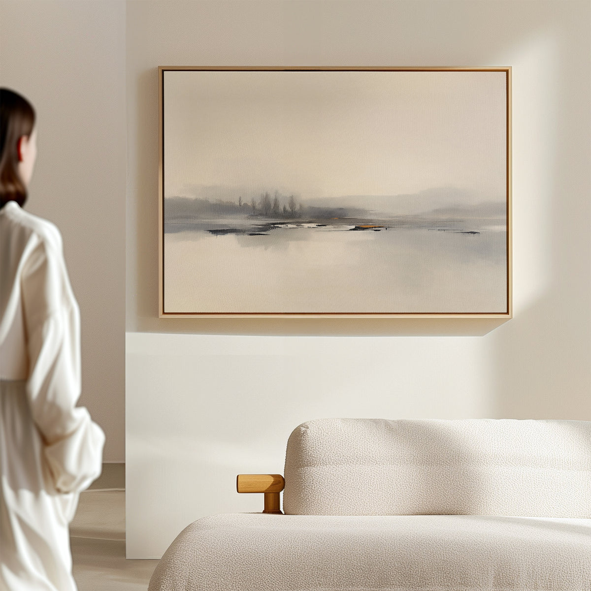 A person stands in a softly lit room, gazing at the "Peaceful Lake Morning Canvas Art - Horizontal" with muted gray and beige tones, hanging above a light-colored upholstered sofa and creating a tranquil atmosphere.