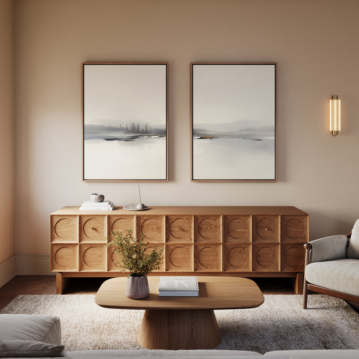 A cozy living room features a wooden sideboard, modern coffee table, light armchair, and the Peaceful Lake Morning - Set Of 2 prints on a beige wall. A small plant and books decorate the table; soft lighting enhances the peaceful vibe.