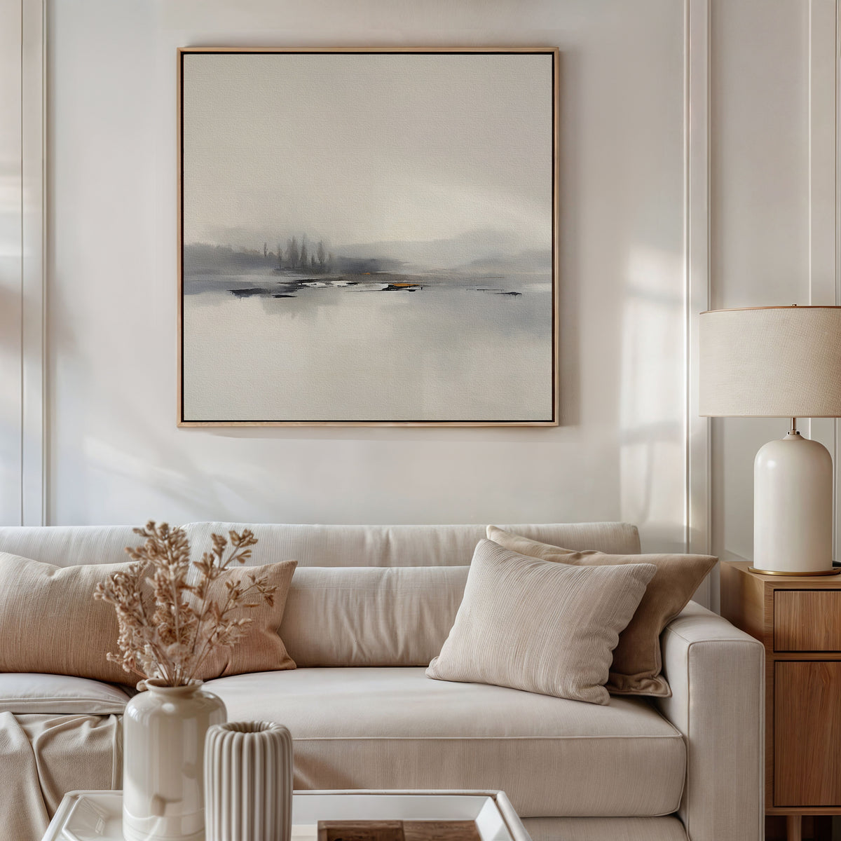 A neutral-toned living room features a beige sofa with matching pillows, a side table with a ceramic lamp, dried flowers in a vase, and the “Peaceful Lake Morning Canvas Art - Square” above the sofa to enhance the tranquil ambiance.