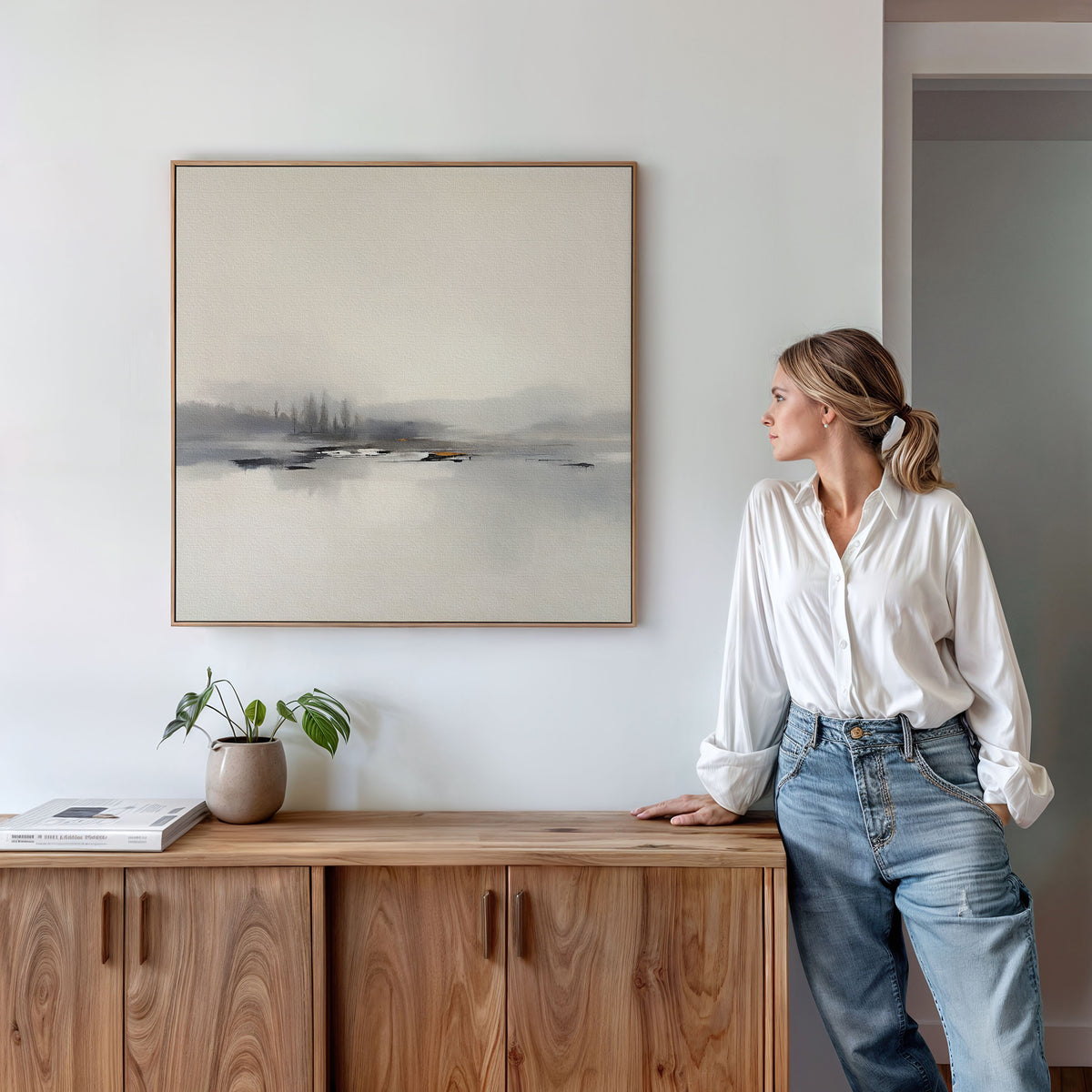 A woman in a white blouse and jeans stands by a wooden sideboard, admiring the Peaceful Lake Morning Canvas Art - Square on the wall. A potted plant and a book add to the tranquil feel of this calm, modern space.