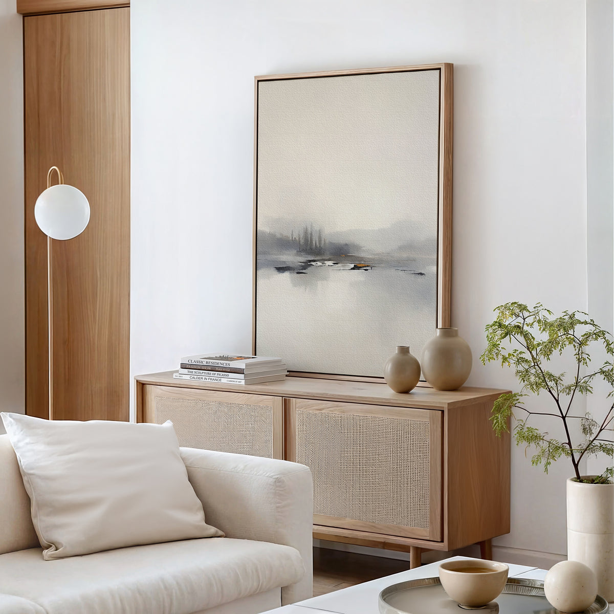 A modern, minimalist living room featuring a light wood sideboard, the Peaceful Lake Morning Canvas Art - Vertical, ceramic vases, a small plant, and a white sofa with cushion. Neutral tones and natural textures create a calm atmosphere.