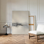 A serene, minimalist room with a white cushioned chair, a small round black table with books and candles, and the Peaceful Lake Morning Canvas Art - Vertical resting against a paneled white wall on herringbone wood floors.
