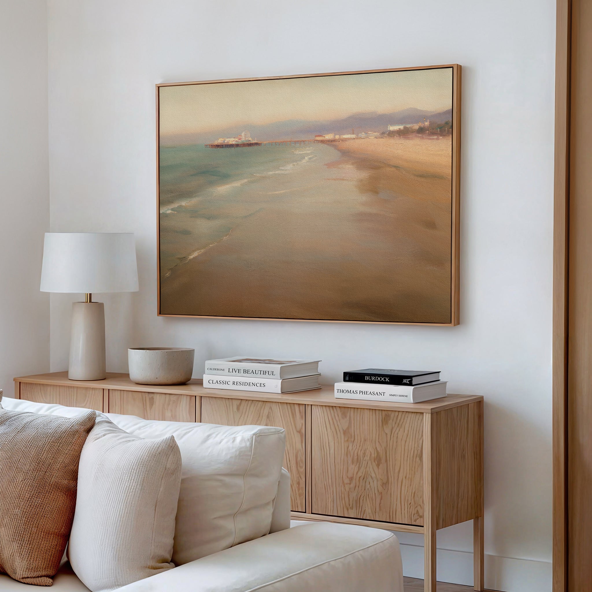 A modern living room with a beige sofa, light wood sideboard, stacked books, white lamp, bowl, and dreamy "Pier Dreams Canvas Art - Horizontal" beach scene displayed on the wall above the sideboard.