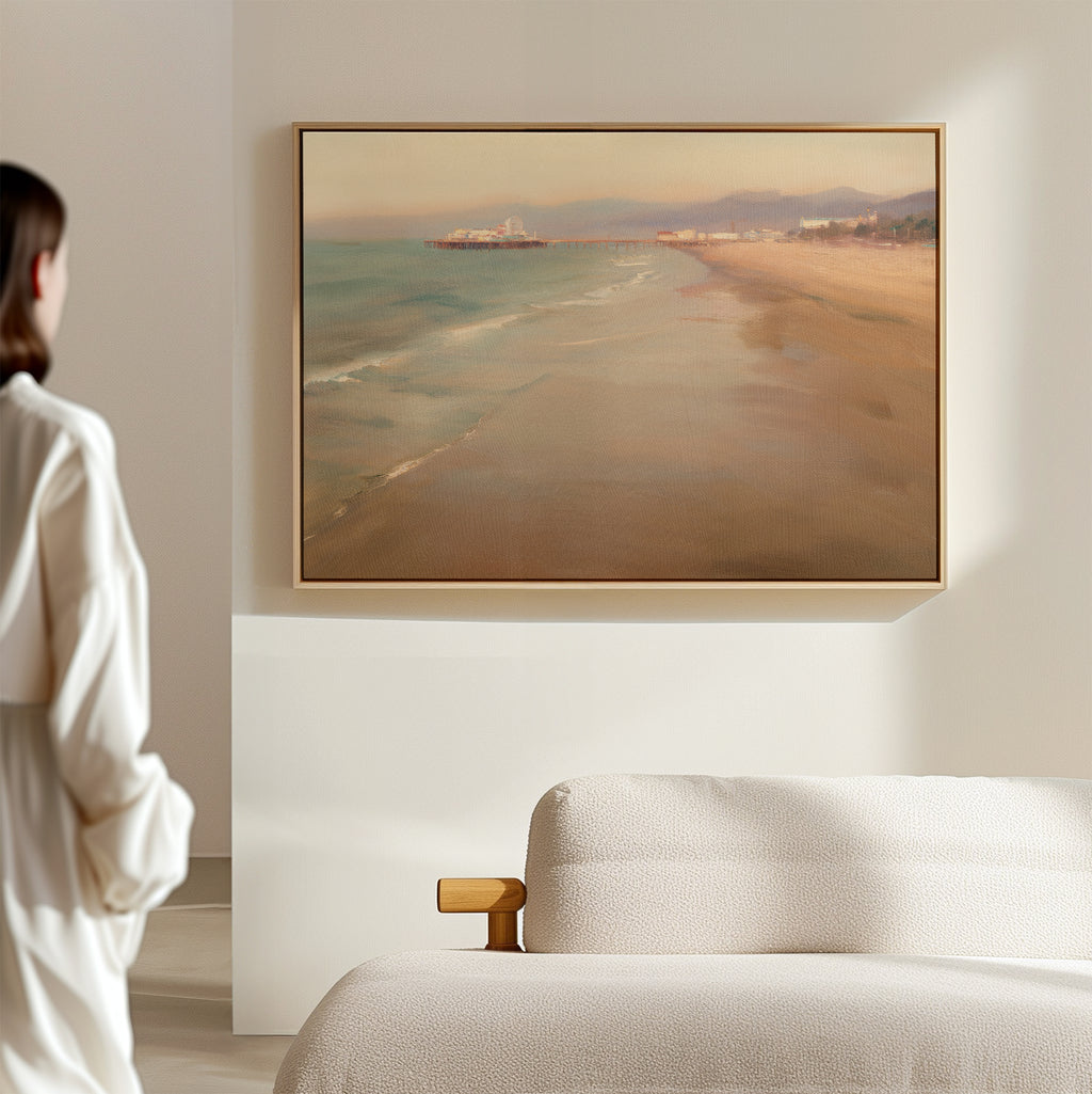 A woman in a white robe stands in a minimalist room, gazing at Pier Dreams Canvas Art - Horizontal, a framed painting of a serene beach and pier, hanging above a light-colored textured sofa.