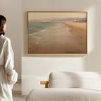 A woman in a white robe stands in a minimalist room, gazing at Pier Dreams Canvas Art - Horizontal, a framed painting of a serene beach and pier, hanging above a light-colored textured sofa.