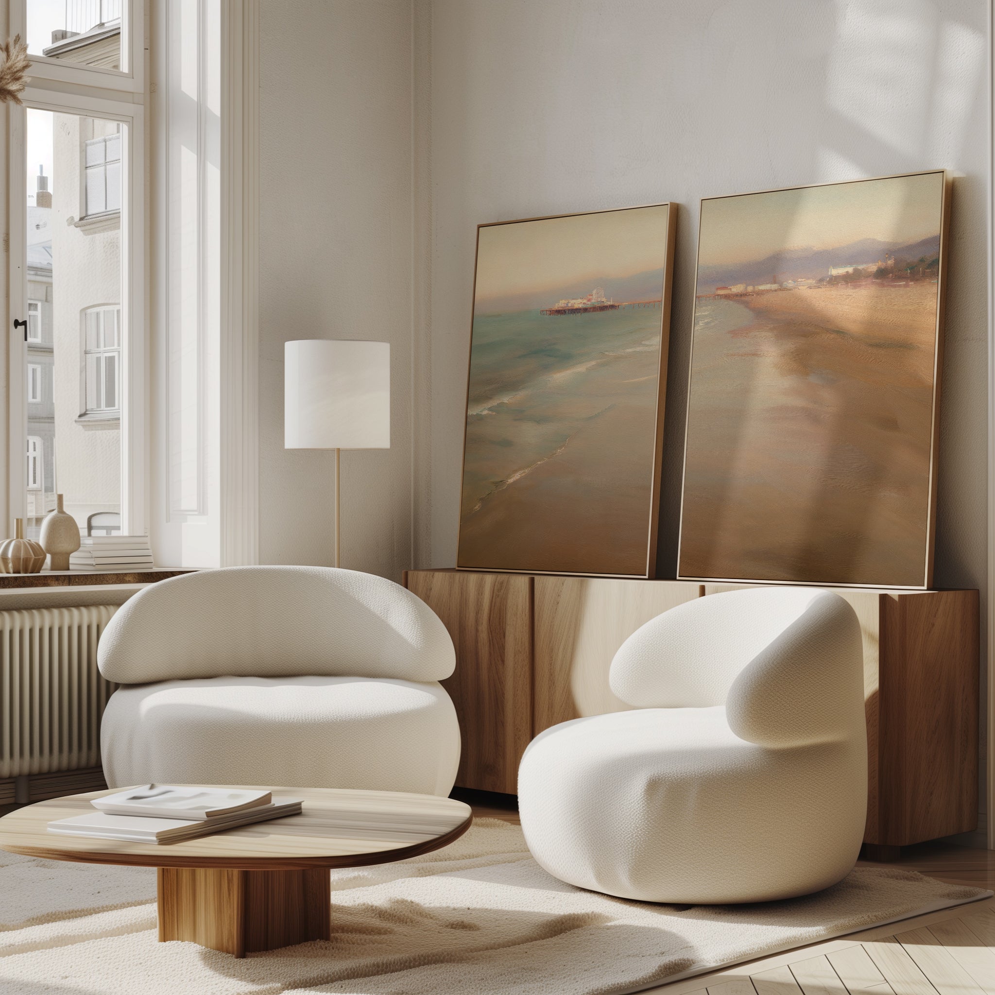 A bright, modern living room with two white curved armchairs, a low wooden coffee table, and Pier Dreams - Set of 2 large framed beach landscapes leaning against a light wood sideboard by a tall window.