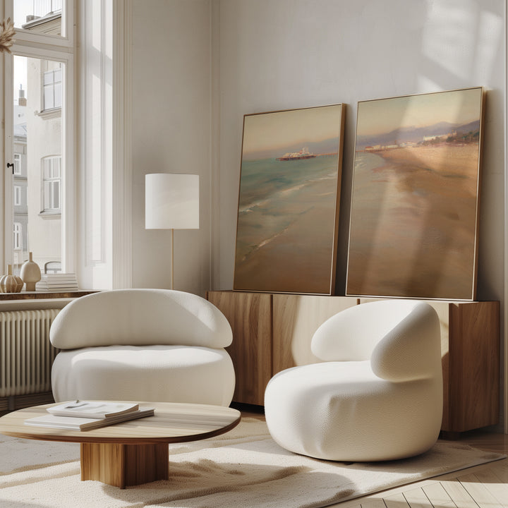 A bright, modern living room with two white curved armchairs, a low wooden coffee table, and Pier Dreams - Set of 2 large framed beach landscapes leaning against a light wood sideboard by a tall window.