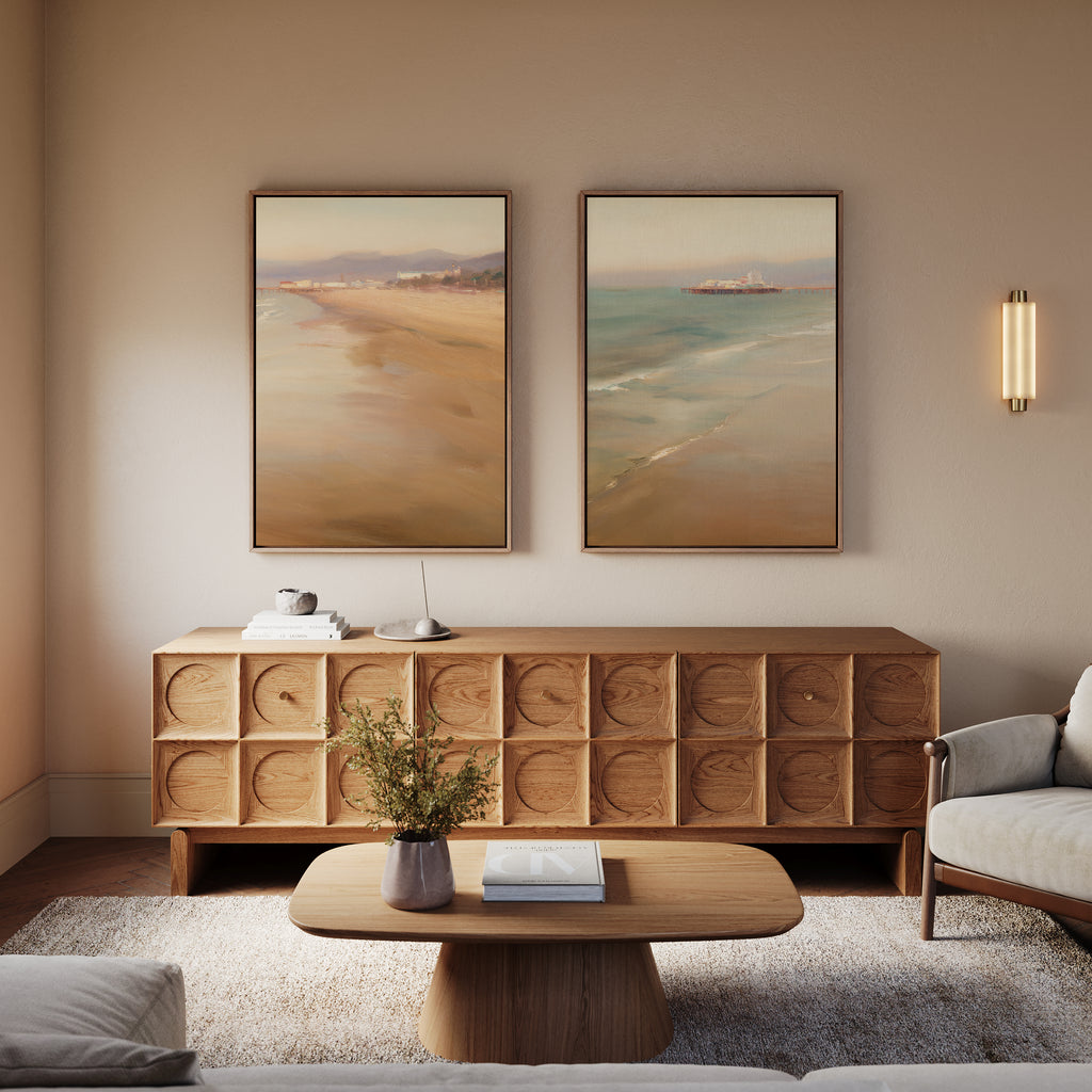 A cozy living room with a modern wooden sideboard, a wooden coffee table with a book and vase, light armchairs, and “Pier Dreams - Set of 2” dreamy beach landscape wall art above the sideboard. Warm lighting adds to the calm atmosphere.