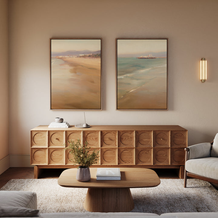 A cozy living room with a modern wooden sideboard, a wooden coffee table with a book and vase, light armchairs, and “Pier Dreams - Set of 2” dreamy beach landscape wall art above the sideboard. Warm lighting adds to the calm atmosphere.