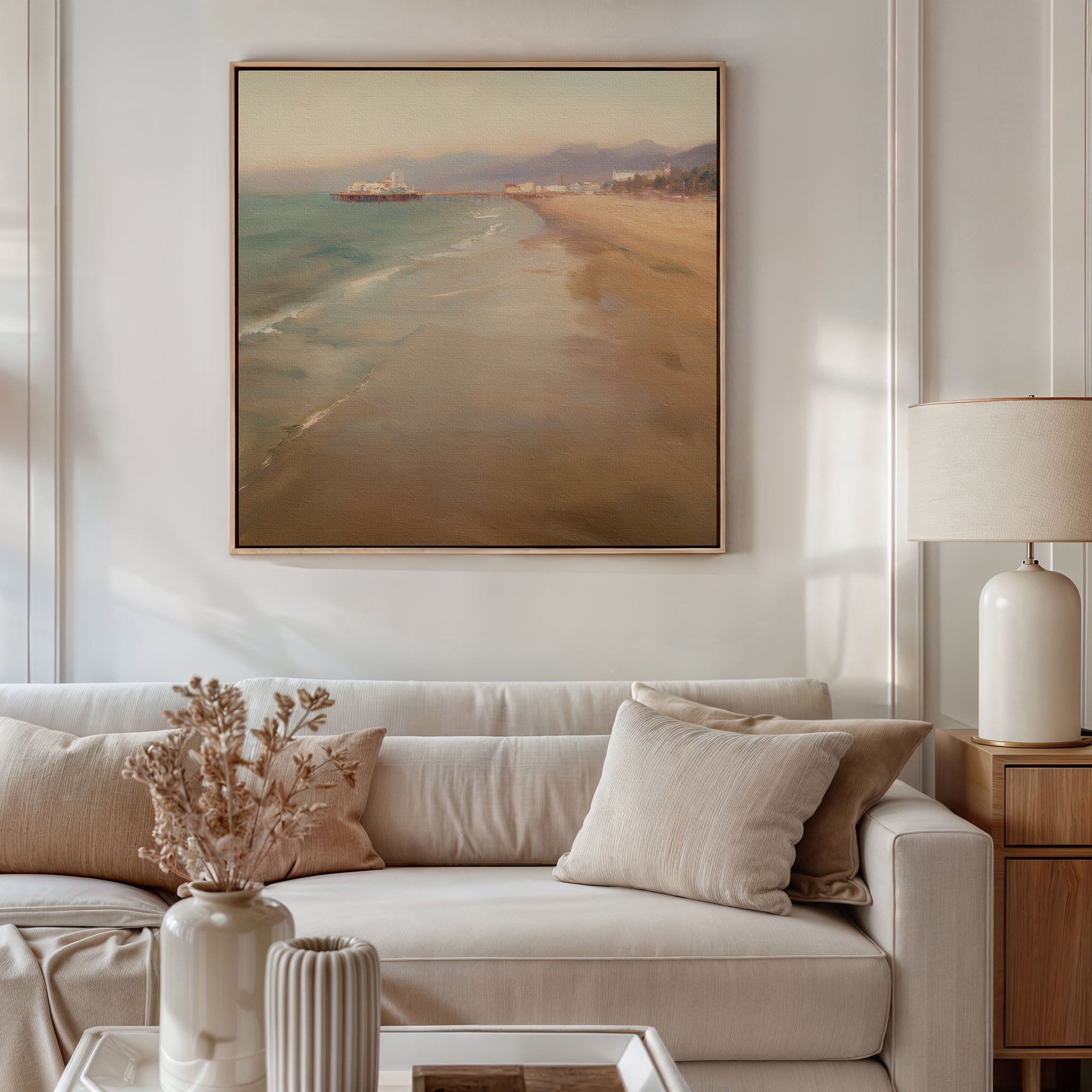 A cozy living room with a white sofa, beige pillows, a side table with a white lamp, and a vase of dried flowers. Above the sofa hangs the Pier Dreams Canvas Art - Square, adding coastal charm to the space.