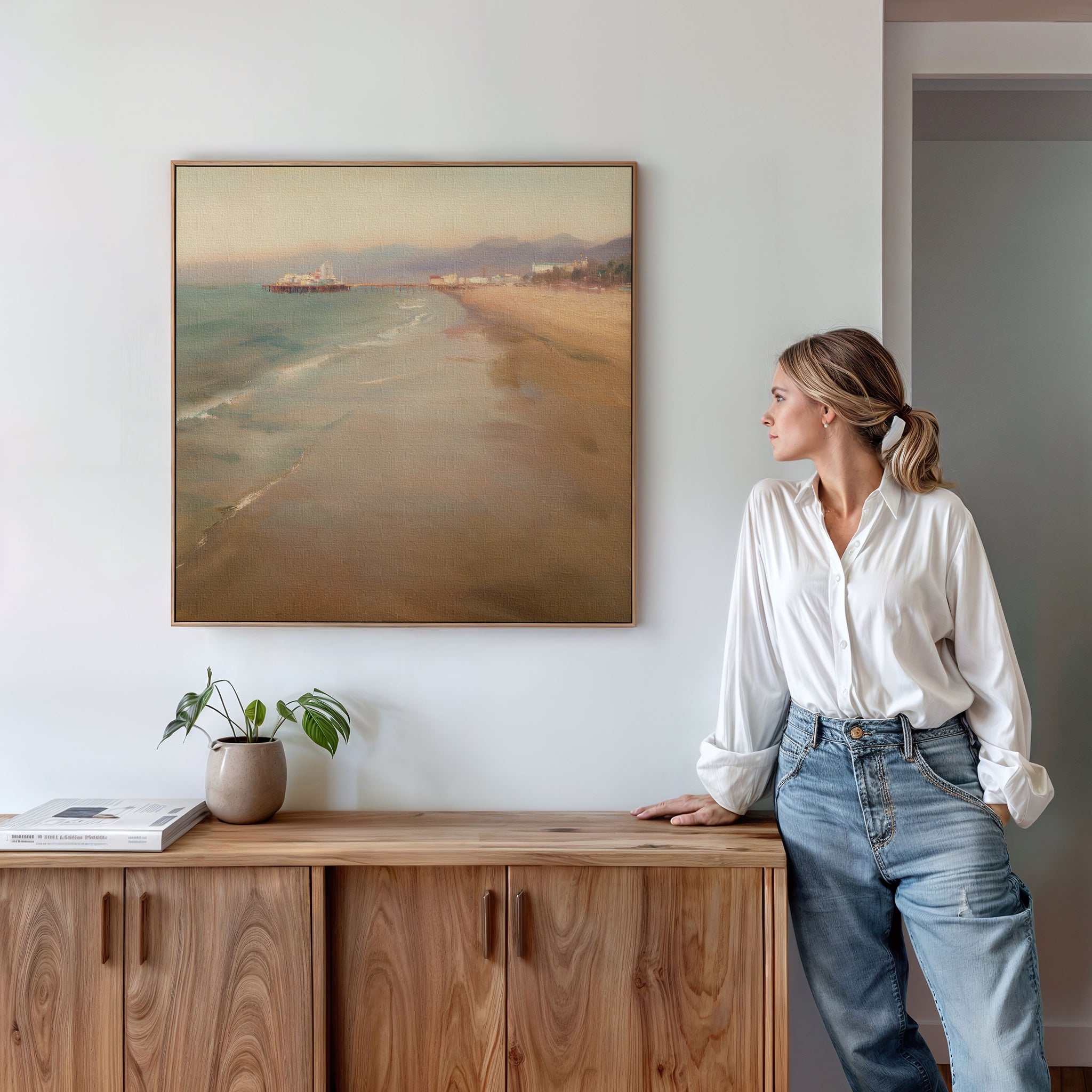 A woman in a white blouse and jeans admires the Pier Dreams Canvas Art - Square, featuring a coastal beach scene on a white wall above a wooden sideboard with a small plant and book.
