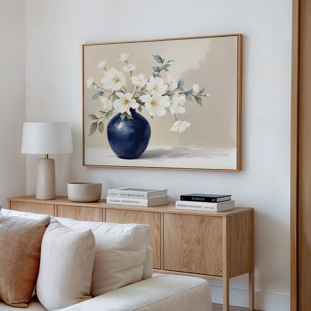 A minimalist living room corner featuring a wooden sideboard, stacked books, a lamp, bowl, and the Porcelain Dreams Canvas Art - Horizontal—an elegant botanical print of white flowers in a blue vase—hanging above.