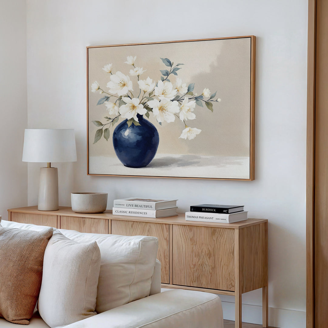 A minimalist living room corner featuring a wooden sideboard, stacked books, a lamp, bowl, and the Porcelain Dreams Canvas Art - Horizontal—an elegant botanical print of white flowers in a blue vase—hanging above.