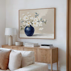 A minimalist living room corner featuring a wooden sideboard, stacked books, a lamp, bowl, and the Porcelain Dreams Canvas Art - Horizontal—an elegant botanical print of white flowers in a blue vase—hanging above.