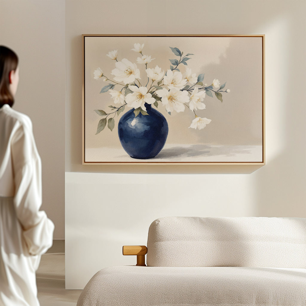 A person stands in a softly lit room, admiring the Porcelain Dreams Canvas Art - Horizontal, which features white flowers in a blue vase. The calm, minimalist decor and beige sofa highlight its Nordic Park-inspired elegance and floral beauty.
