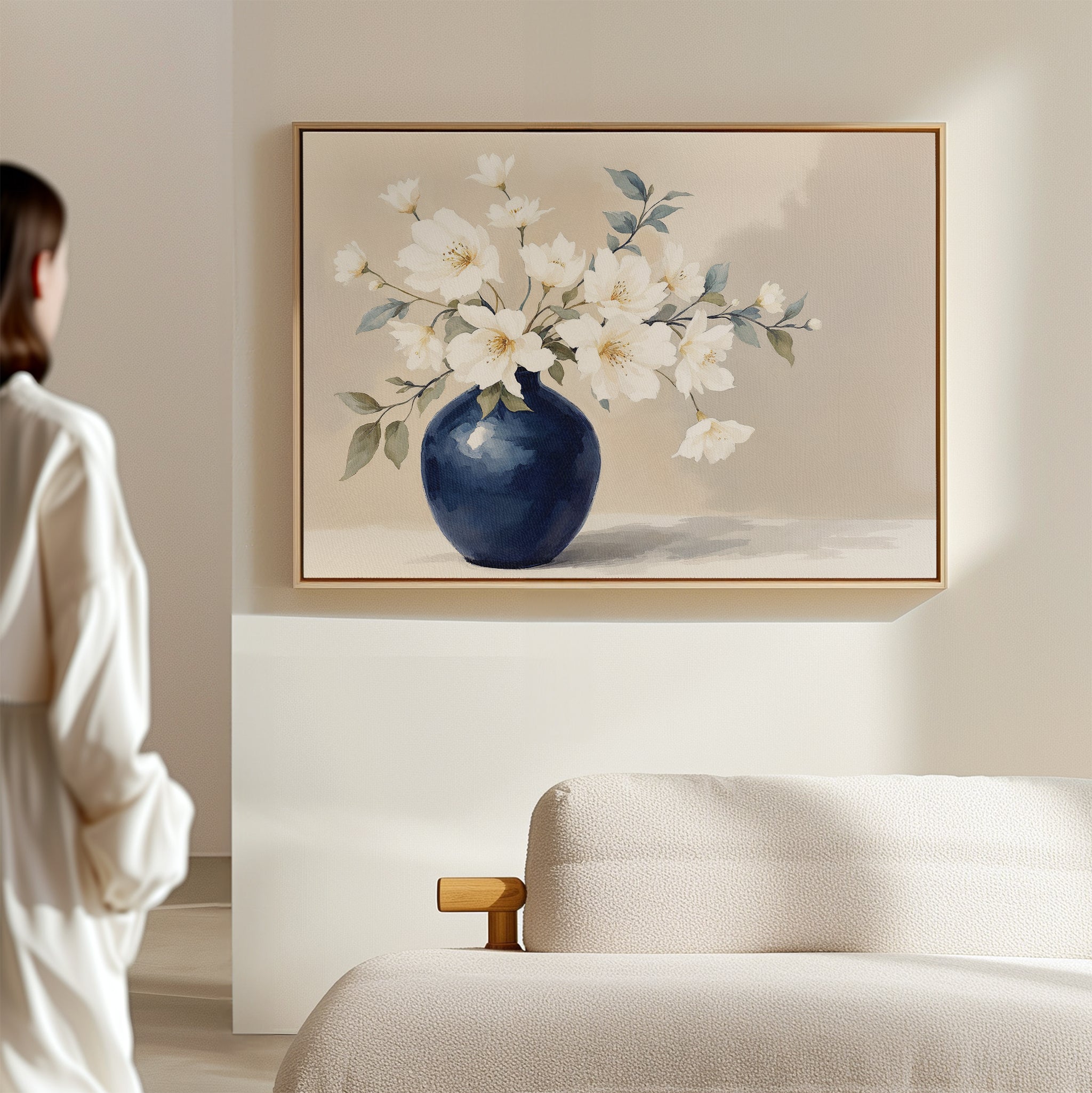 A person stands in a softly lit room, admiring the Porcelain Dreams Canvas Art - Horizontal, which features white flowers in a blue vase. The calm, minimalist decor and beige sofa highlight its Nordic Park-inspired elegance and floral beauty.