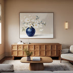 A modern living room featuring the Porcelain Dreams Canvas Art - Horizontal depicting white flowers in a blue vase, a wooden sideboard, light wood coffee table with flowers, sofa, neutral walls, and a wall sconce.