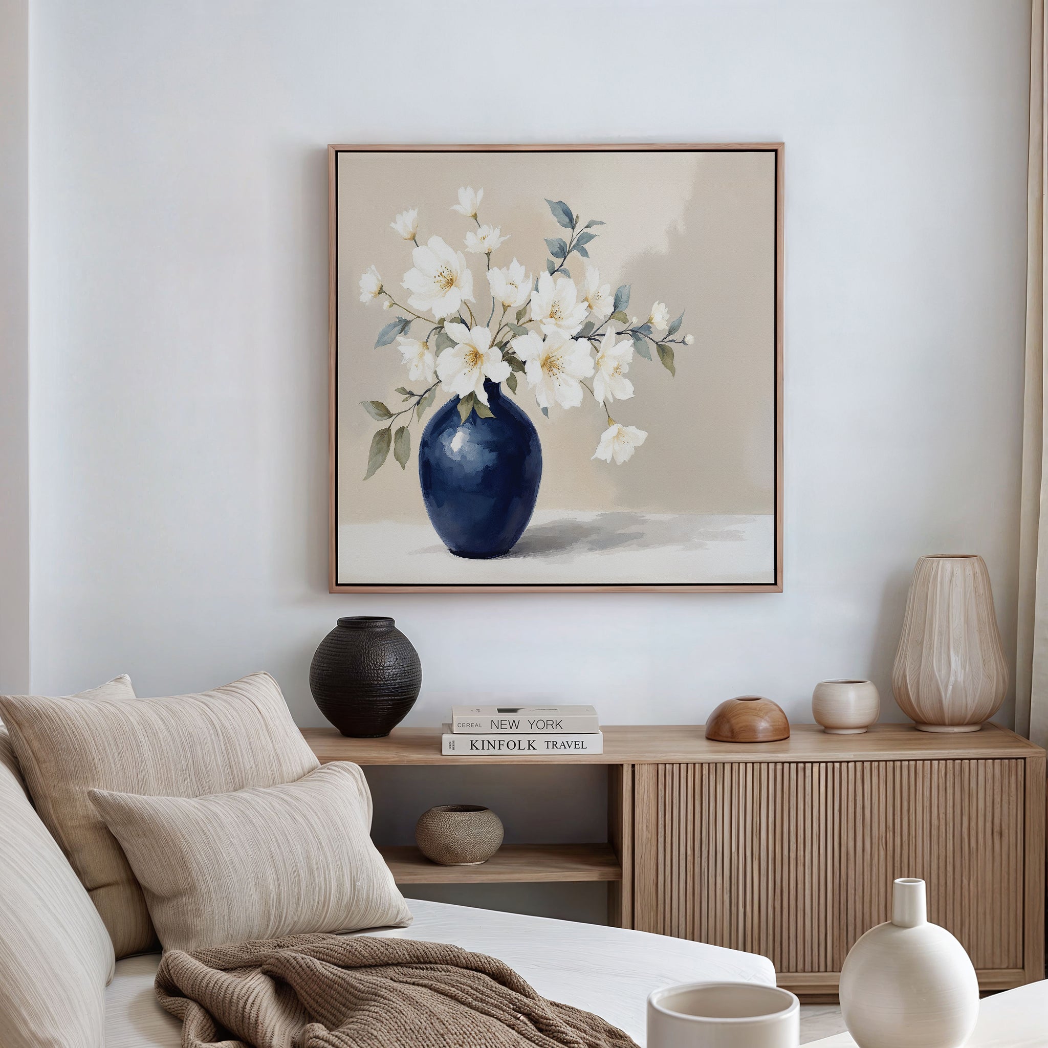 A modern, neutral-toned living room features a beige sofa with throw pillows, a wooden sideboard with vases and books, and the Porcelain Dreams Canvas Art - Square painting of white flowers in a navy blue vase hanging on the wall.
