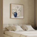A minimalist bedroom with a beige upholstered bed, white pillows, and the Porcelain Dreams Canvas Art - Square, featuring white flowers in a navy blue vase above the headboard. Soft lighting and neutral tones create a calm atmosphere.