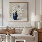 A neutral living room with a beige sofa, cream cushions, and a side table with a lamp. Above the sofa hangs the Porcelain Dreams Canvas Art - Square, showing white flowers in a navy vase; dried flowers decorate the coffee table.