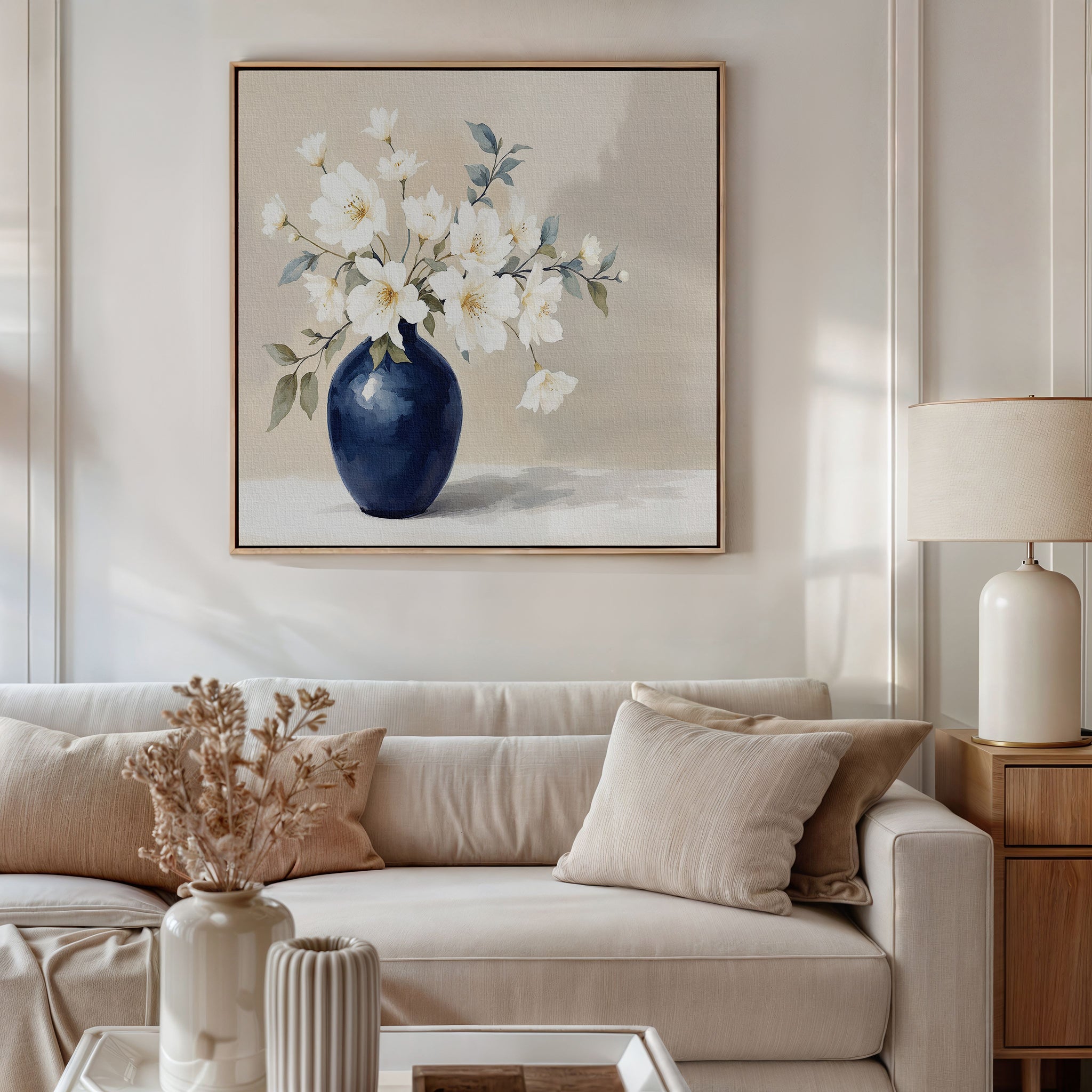 A neutral living room with a beige sofa, cream cushions, and a side table with a lamp. Above the sofa hangs the Porcelain Dreams Canvas Art - Square, showing white flowers in a navy vase; dried flowers decorate the coffee table.