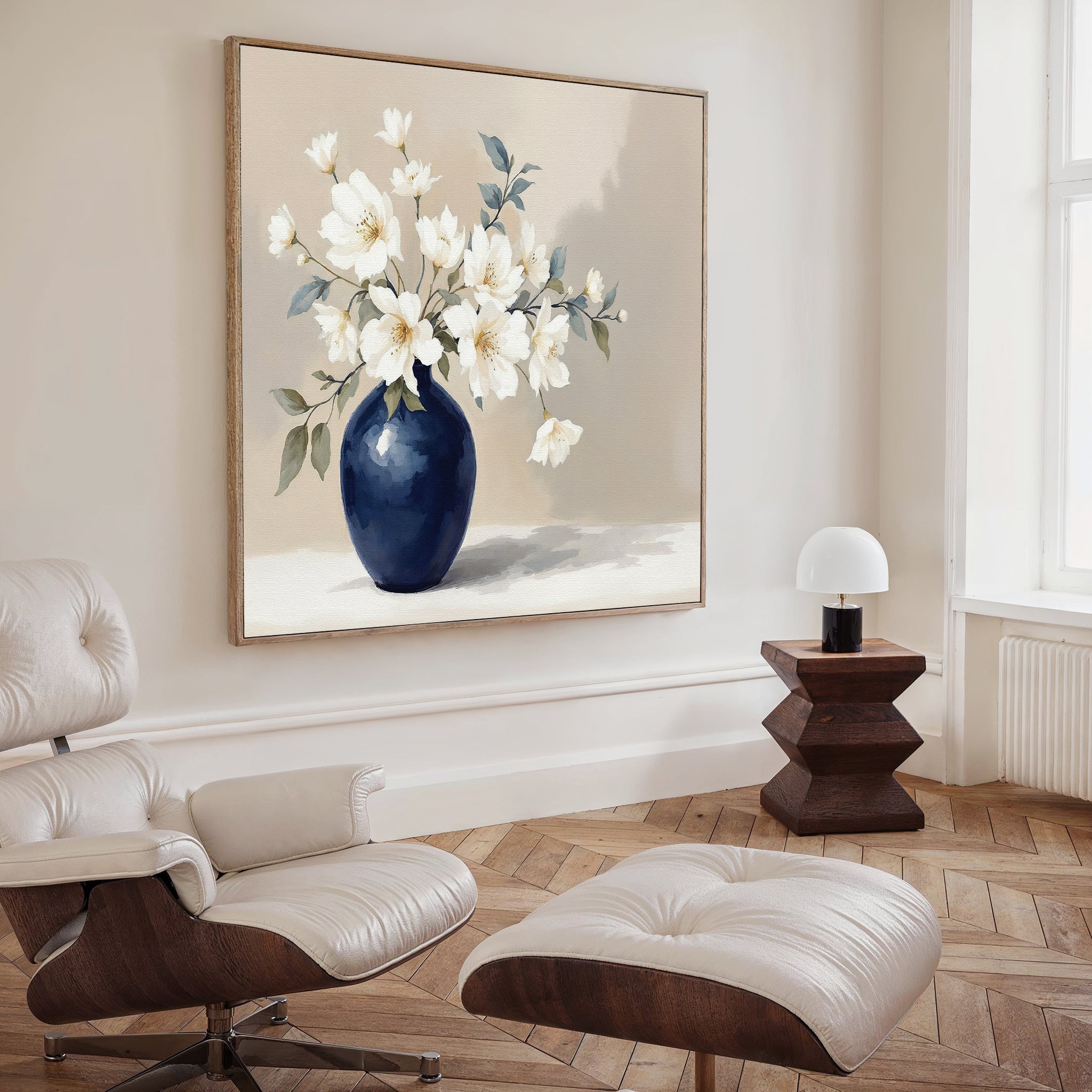 A modern living room with a white lounge chair and ottoman, a dark wood side table with a white lamp, and the Porcelain Dreams Canvas Art - Square featuring white flowers in a navy blue vase on the wall.