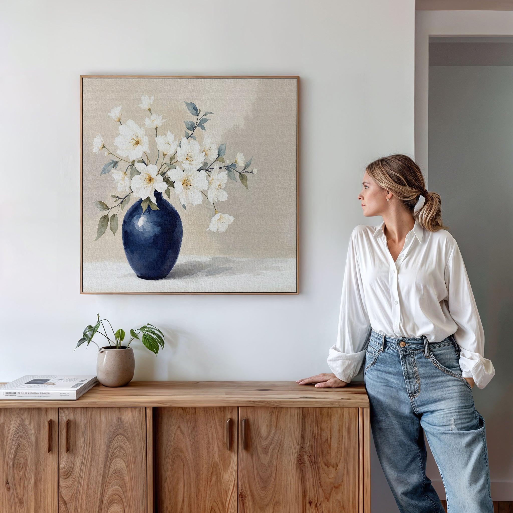 A woman in a white blouse and jeans leans against a wooden cabinet, looking at the Porcelain Dreams Canvas Art - Square featuring white flowers in a navy blue vase above her. A small plant and book sit on the cabinet.