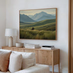 A cozy living room with a light wood sideboard, lamp, books, and a bowl. Above the sideboard hangs the Prairie Wanderer Canvas Art - Horizontal, showing a person on horseback in grassy hills with mountains in the background.
