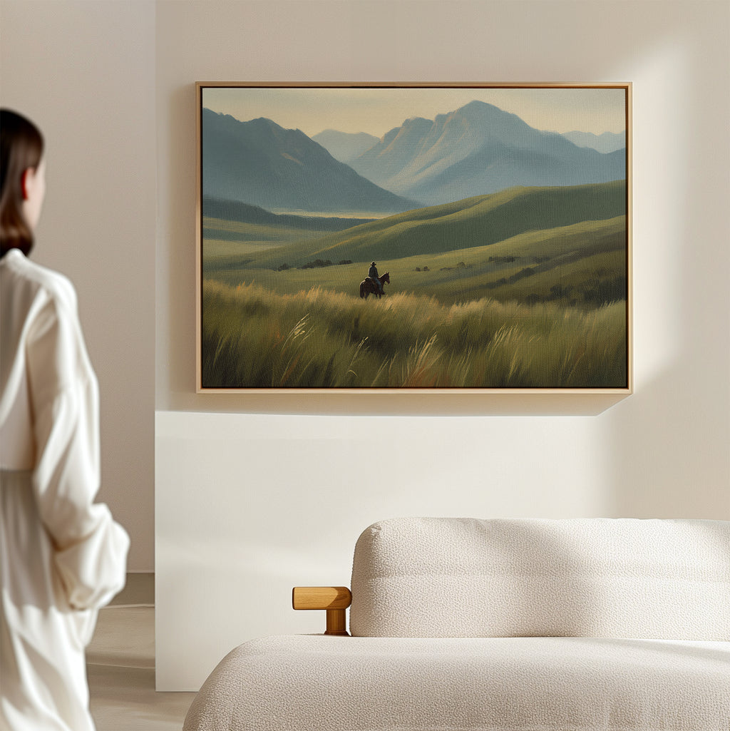 A woman stands in a minimalist room, admiring the Prairie Wanderer Canvas Art - Horizontal featuring a lone horseback rider in a grassy valley, framed above a beige bed—ideal prairie decor for serene spaces.