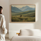 A woman stands in a minimalist room, admiring the Prairie Wanderer Canvas Art - Horizontal featuring a lone horseback rider in a grassy valley, framed above a beige bed—ideal prairie decor for serene spaces.