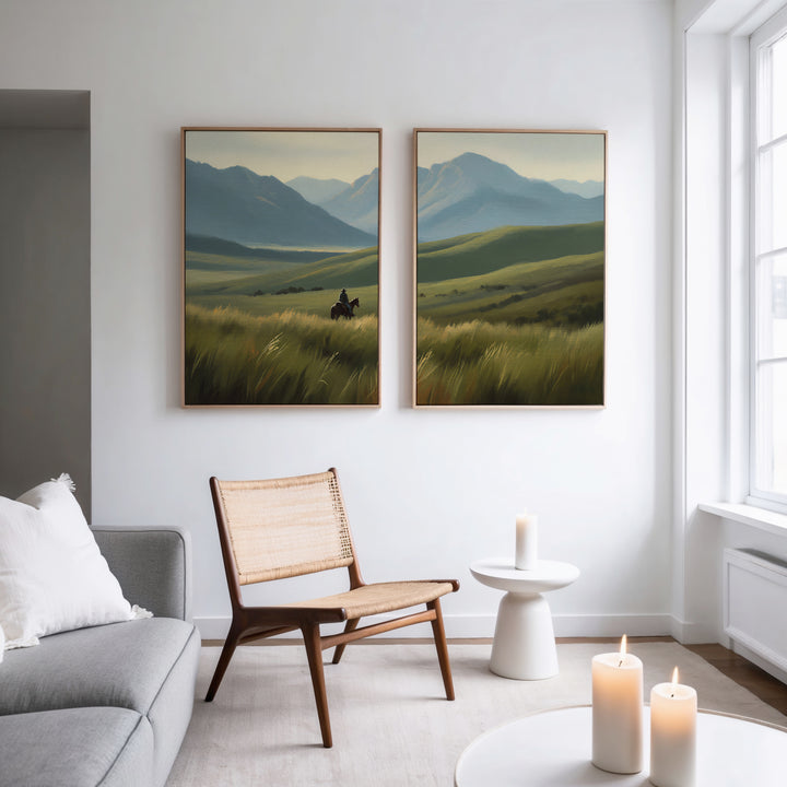 A modern living room features the Prairie Wanderer - Set of 2 mountain canvas prints of grassy fields, hung above a wooden chair, side table, and candles. Light from large windows enhances the cozy, minimalist atmosphere.