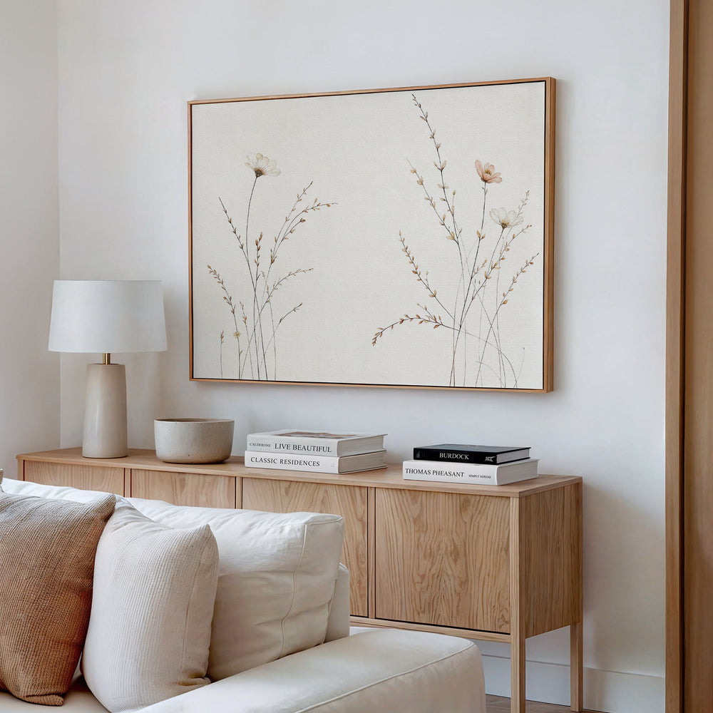 A modern living room corner with a light wood sideboard, white lamp, decorative bowl, stacked books, beige sofa, and the Prairie Whispers Canvas Art - Horizontal featuring delicate neutral-toned flowers and grasses for natural tranquility.