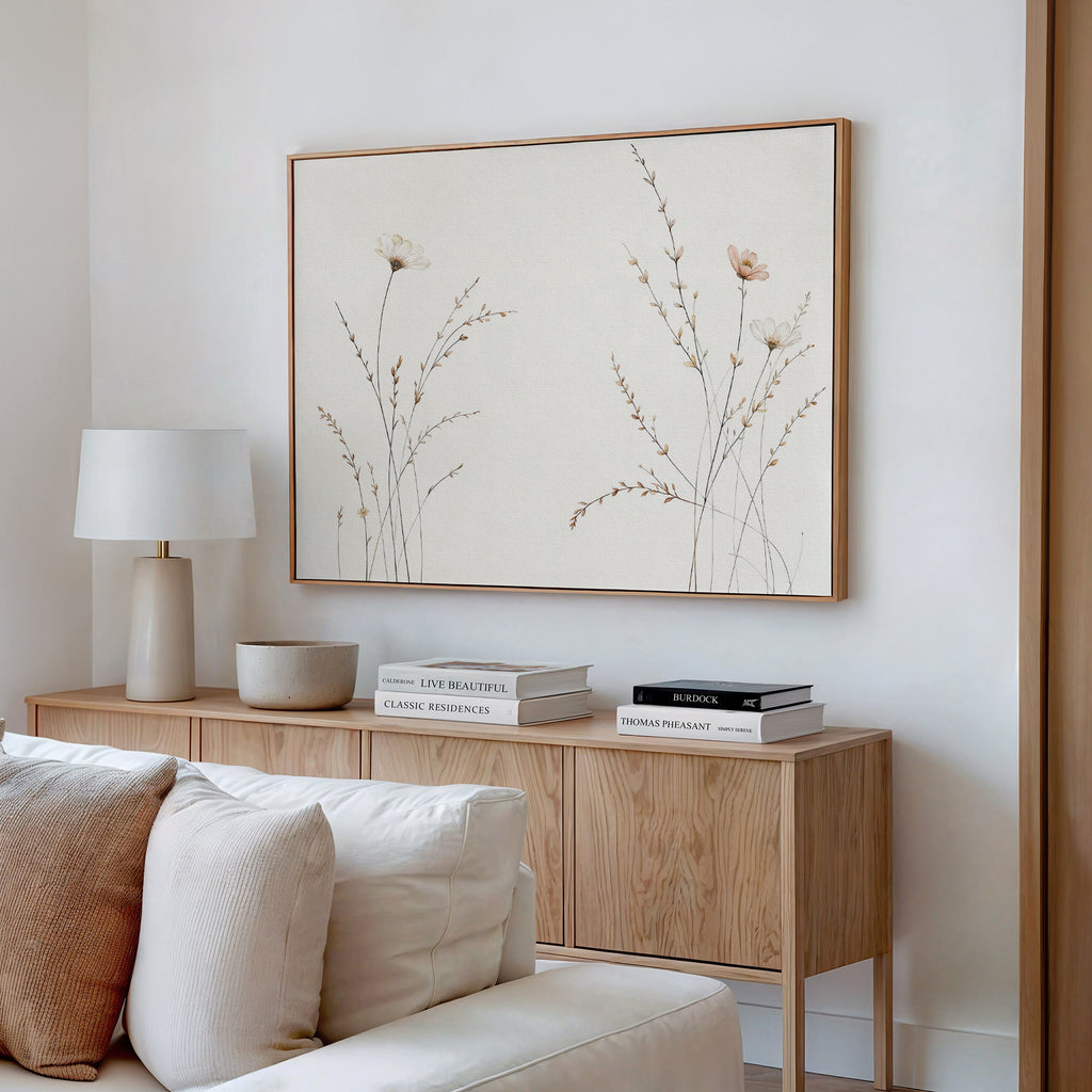 A modern living room corner with a light wood sideboard, white lamp, decorative bowl, stacked books, beige sofa, and the Prairie Whispers Canvas Art - Horizontal featuring delicate neutral-toned flowers and grasses for natural tranquility.