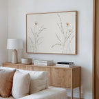 A modern living room corner with a light wood sideboard, white lamp, decorative bowl, stacked books, beige sofa, and the Prairie Whispers Canvas Art - Horizontal featuring delicate neutral-toned flowers and grasses for natural tranquility.