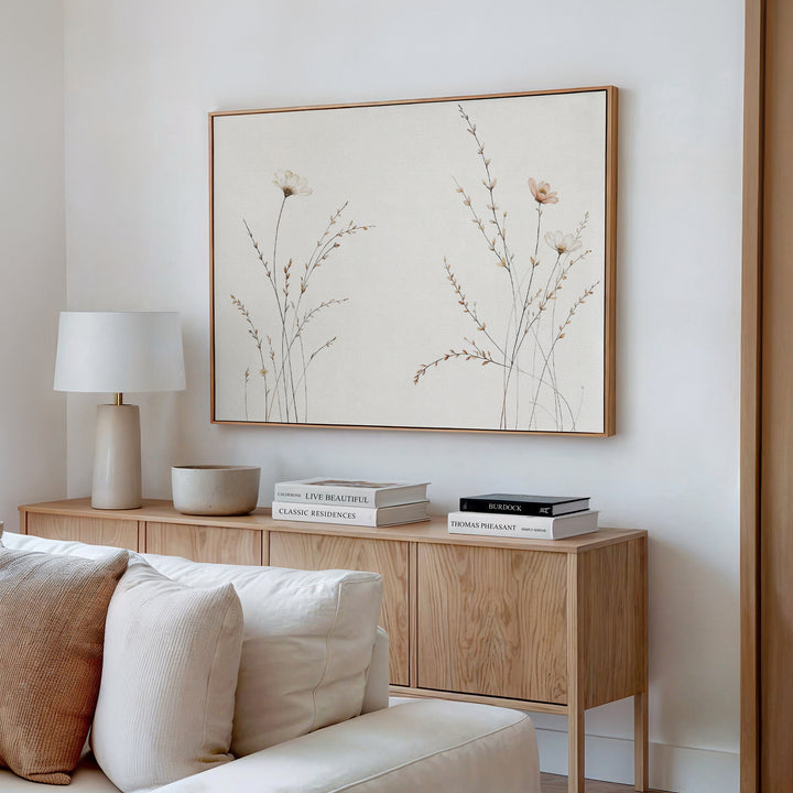 A modern living room corner with a light wood sideboard, white lamp, decorative bowl, stacked books, beige sofa, and the Prairie Whispers Canvas Art - Horizontal featuring delicate neutral-toned flowers and grasses for natural tranquility.