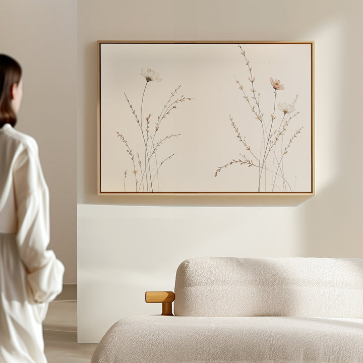 Dressed in white, a person stands by a beige sofa, admiring the Prairie Whispers Canvas Art - Horizontal with delicate wildflowers on a minimalist light wall, creating a modern living room of natural tranquility.