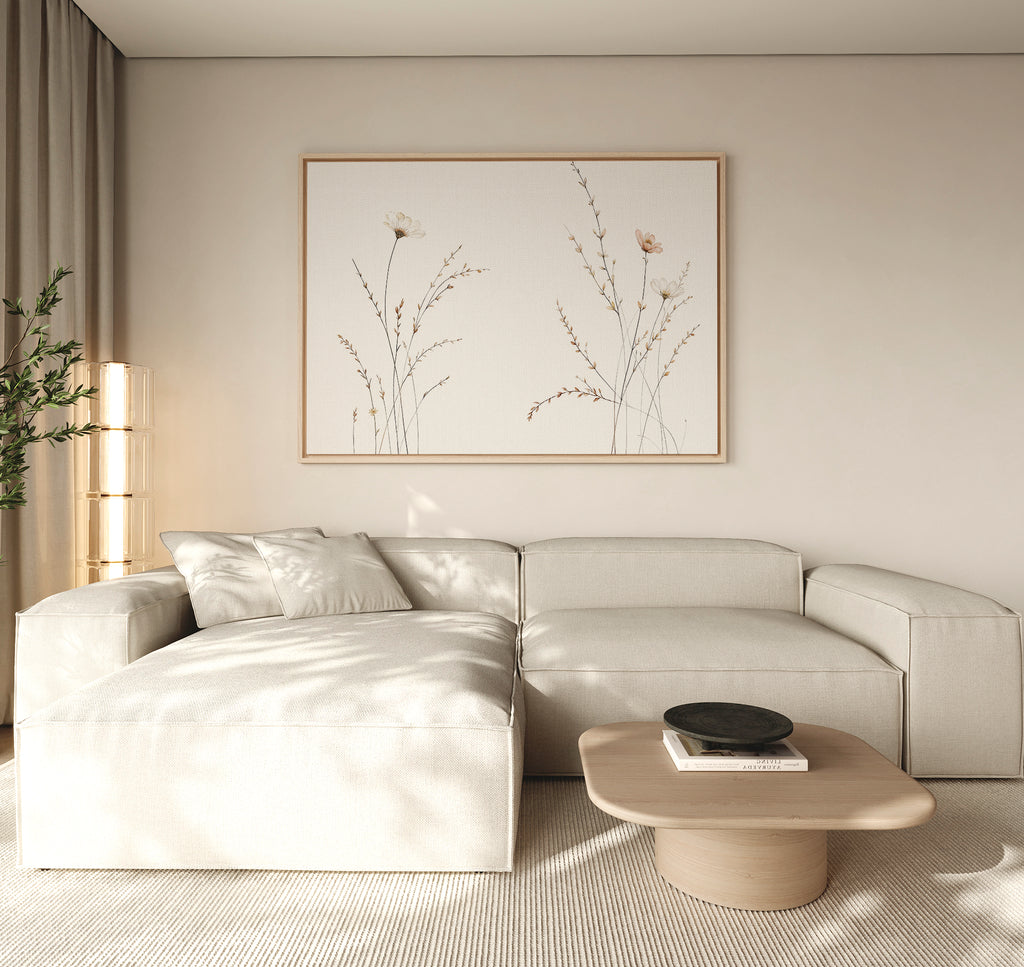 A minimalist living room features a light beige sectional sofa, a low wooden coffee table with a black bowl, neutral walls, and the Prairie Whispers Canvas Art - Horizontal as botanical artwork, bathed in soft natural light for tranquil ambiance.