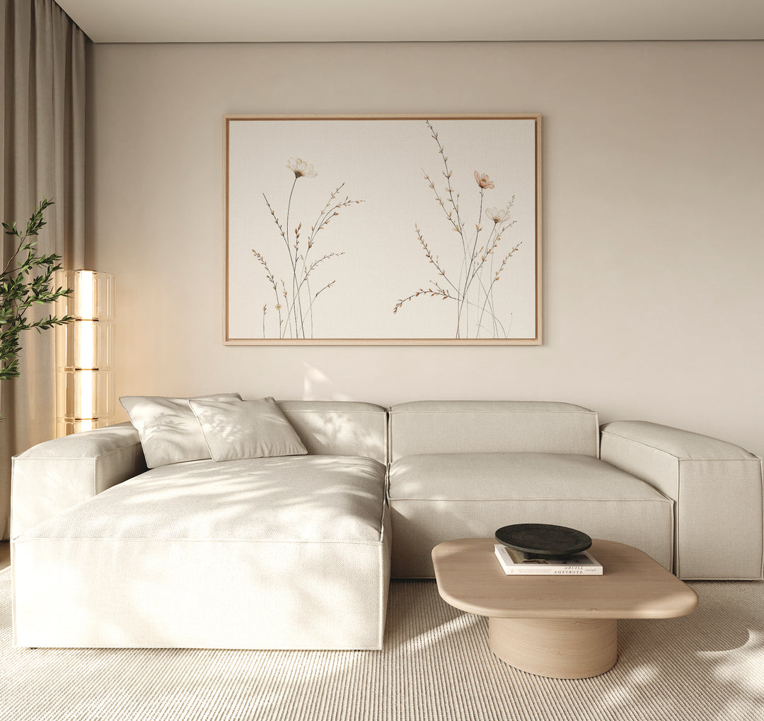 A minimalist living room features a light beige sectional sofa, a low wooden coffee table with a black bowl, neutral walls, and the Prairie Whispers Canvas Art - Horizontal as botanical artwork, bathed in soft natural light for tranquil ambiance.