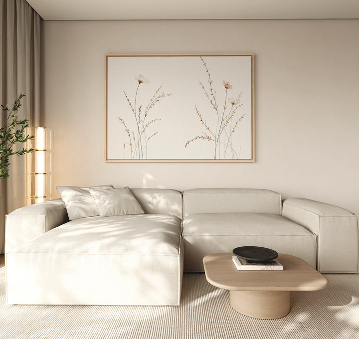 A minimalist living room features a light beige sectional sofa, a low wooden coffee table with a black bowl, neutral walls, and the Prairie Whispers Canvas Art - Horizontal as botanical artwork, bathed in soft natural light for tranquil ambiance.