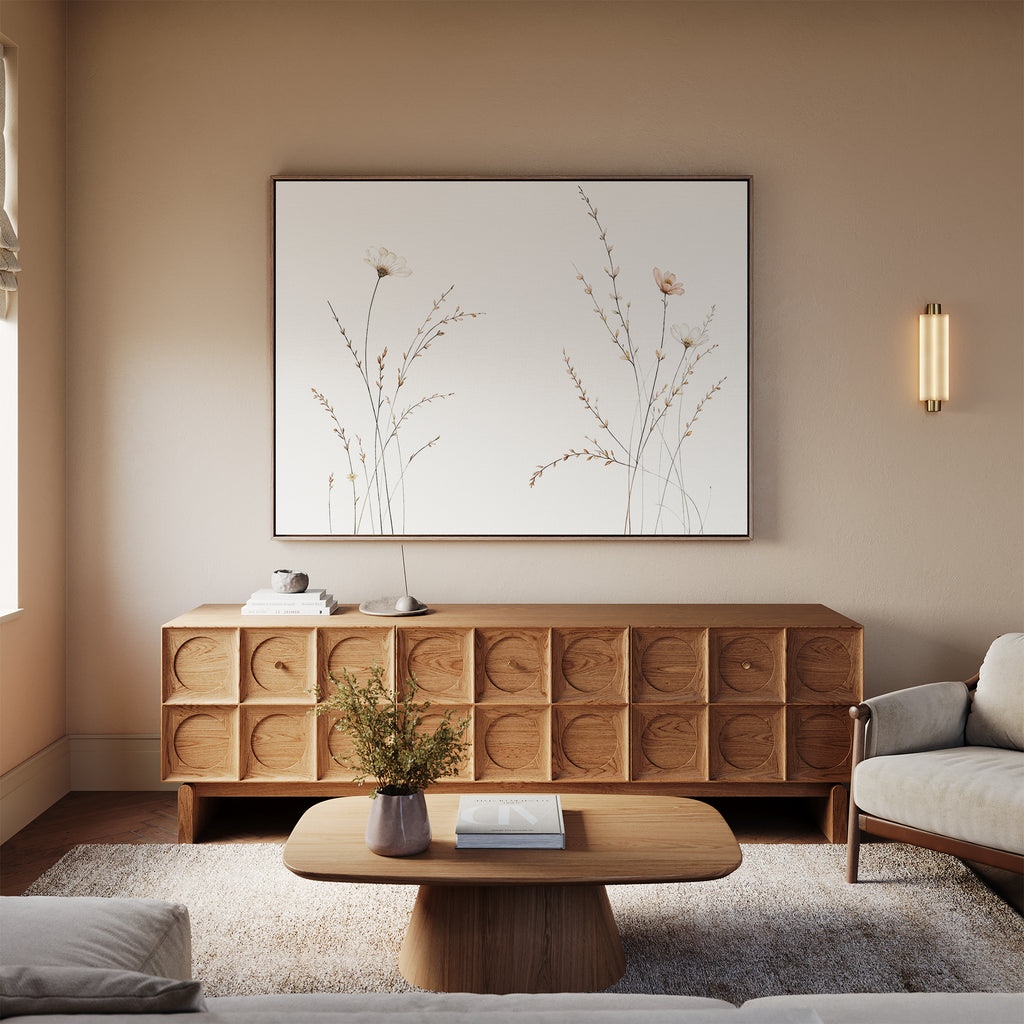 A modern living room features a light wood sideboard, beige sofa, low wooden coffee table, and a vase with flowers. Above the sideboard hangs "Prairie Whispers Canvas Art - Horizontal," adding botanical charm to the tranquil, neutral-toned space.