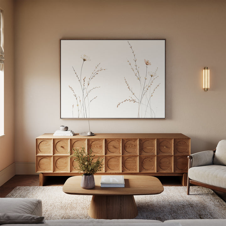 A modern living room features a light wood sideboard, beige sofa, low wooden coffee table, and a vase with flowers. Above the sideboard hangs "Prairie Whispers Canvas Art - Horizontal," adding botanical charm to the tranquil, neutral-toned space.