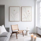 A minimal living room with a wooden chair, round white side table with a lit candle, gray sofa, and Prairie Whispers - Set of 2 botanical wall art on a white wall. Large windows fill the space with natural light.