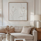 A cozy living room features a white sofa with beige and cream cushions, a wooden side table with a white lamp, and Prairie Whispers Canvas Art - Square on the wall. A vase of dried flowers decorates the coffee table in front.