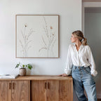 A woman in a white blouse and jeans stands by a wooden cabinet, admiring the Prairie Whispers Canvas Art - Square hanging on a white wall. A small plant and books decorate the cabinet.